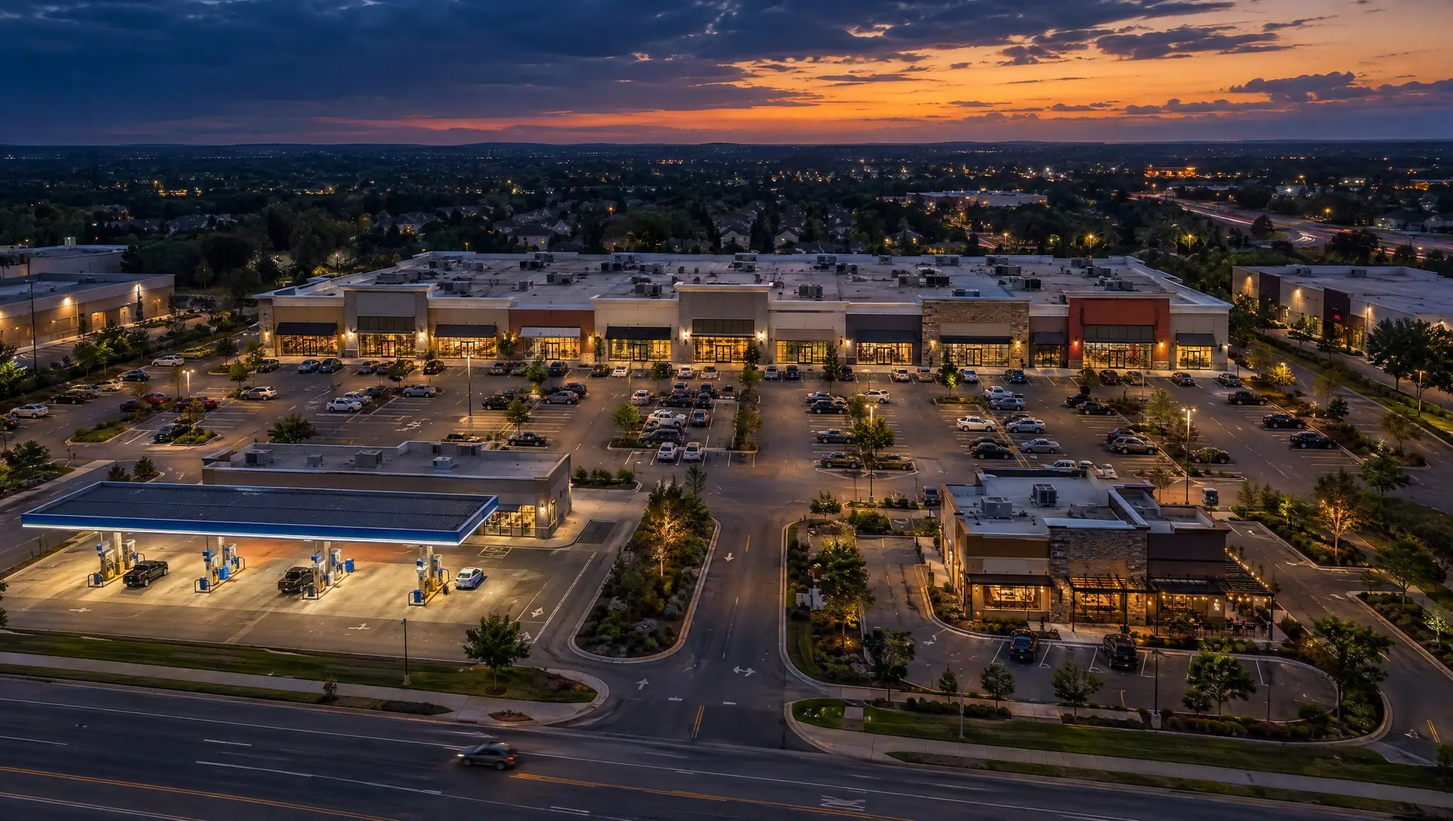 Aerial view of a retail plaza with franchise stores, a fuel station and a parking lot at dusk.